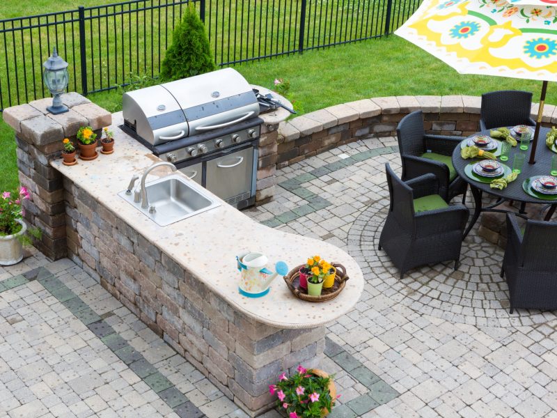 High angle view of a stylish outdoor kitchen, gas barbecue and dining table set for entertaining guests with formal place settings and flowers on a paved patio